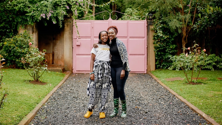 A woman and her daughter stand in front of a pink gate smiling at the camera.