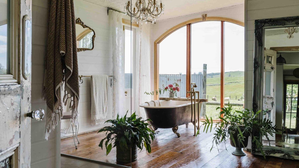 Ein Badezimmer mit einer Klauenfuß-Badewanne vor einem großen Fenster, aus dem man auf ein Feld mit Schafen blickt.