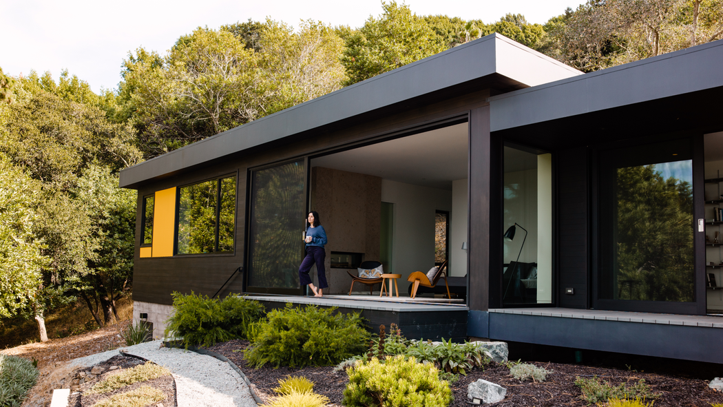 Eine Person steht auf der Terrasse eines großen, modernen, einstöckigen Hauses und blickt auf die Landschaft hinaus.