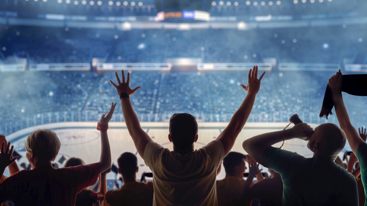 Het silhouet van drie ijshockeyfans, met hun rug naar de camera, terwijl ze juichen in een vol stadion.