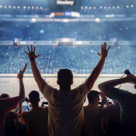 Three hockey fans, with their backs to the camera, are silhouetted by bright lights as they cheer in a crowded arena.