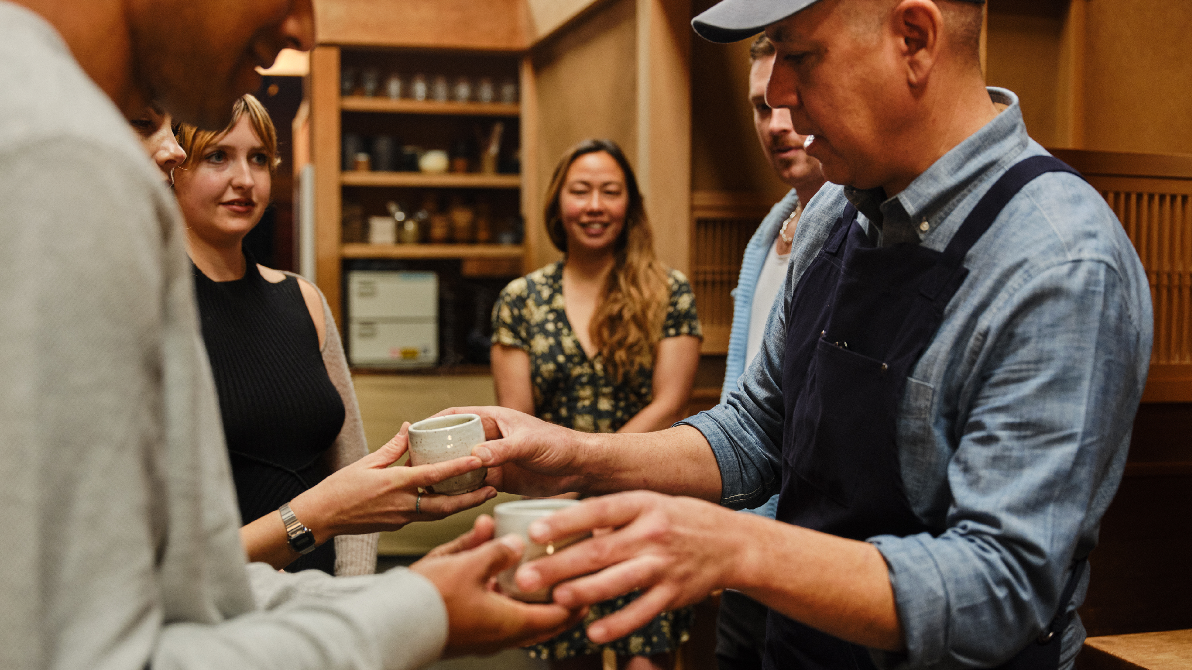 Mtu aliyevaa shati lenye vifungo na aproni ya bluu iliyokolea anatoa vikombe vidogo vya kauri katika chumba kilichopambwa kwa mbao.