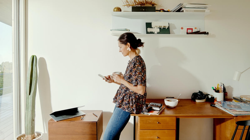 Женщина в джинсах и блузке опирается на стол, держа в одной руке мобильный телефон, а в другой — кружку.