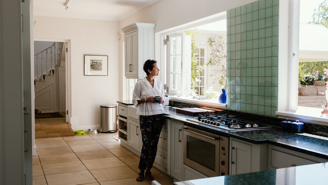 Una persona amb una tassa a la mà està dreta davant d'uns fogons a la cuina i mira per una gran finestra oberta.