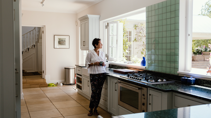 一个拿着咖啡杯的人站在厨房炉灶前,望向敞开的大型窗户外。