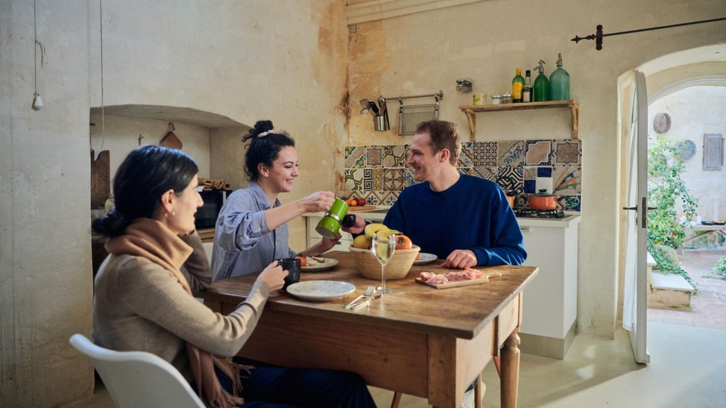 Drei Personen sitzen an einem hölzernen Küchentisch, der für das Frühstück gedeckt ist, mit einem Obstkorb und einem Brett mit Aufschnitt.