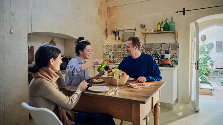 Three people are seated at a wooden kitchen table that’s set for breakfast, with a fruit basket and a charcuterie board.