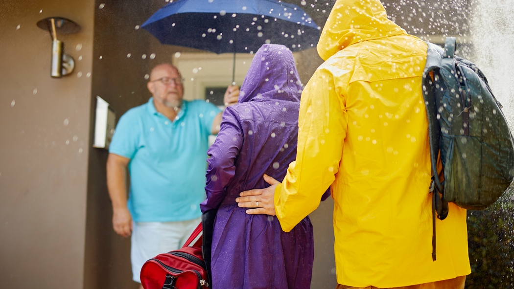 Un hombre sonriente sostiene un paraguas e invita a una pareja a entrar en un alojamiento para que se refugie de la lluvia.