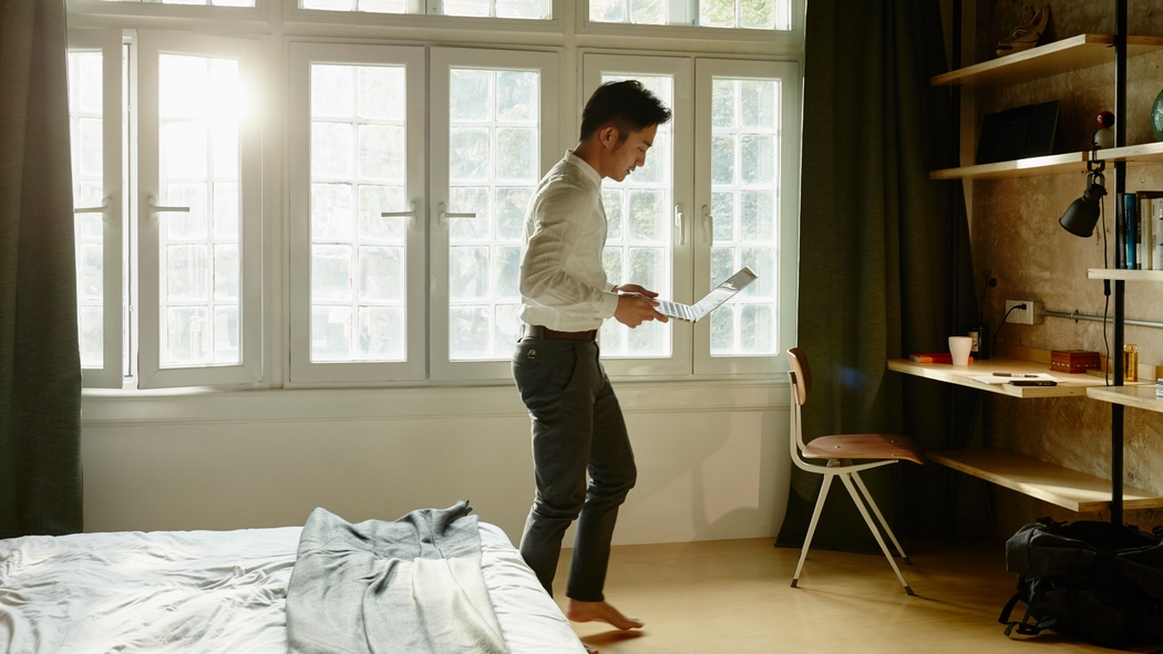A man stands in a sunny bedroom looking at his phone with a large window behind him.