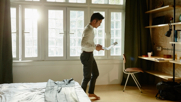A man stands in a sunny bedroom looking at his phone with a large window behind him.