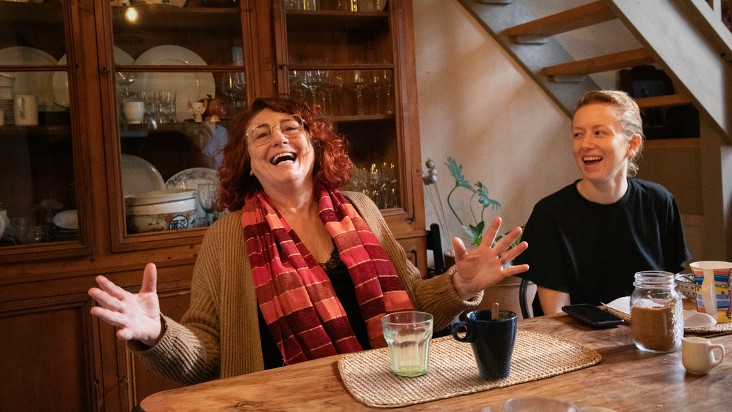 一位出租Airbnb房間的旅居主人與她接待的旅人在一張木桌前並肩而坐，二人正開懷大笑，而桌上則擺放着有耳杯和玻璃杯。
