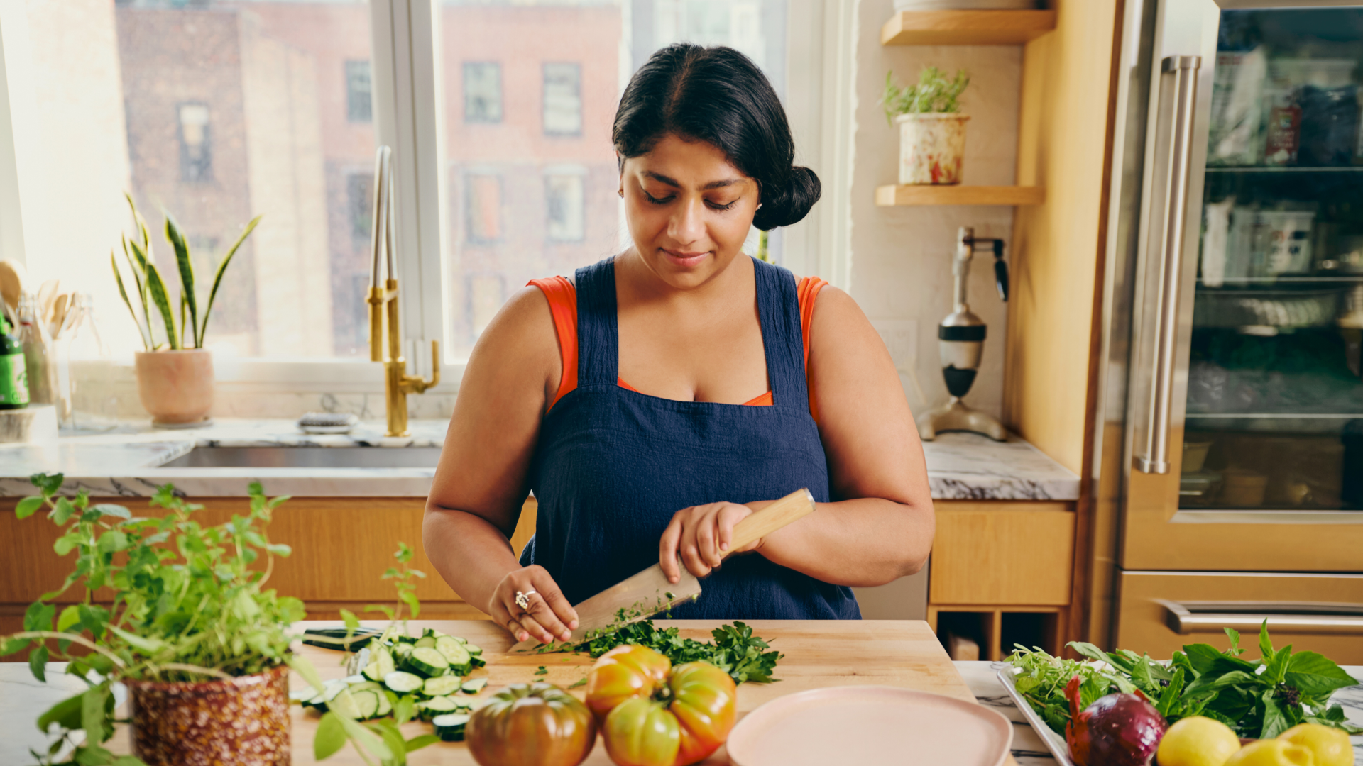 A person chops herbs on a wooden chopping board, with heirloom tomatoes and cucumbers on the board in front of them.