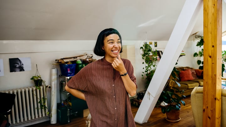 Une personne se tient debout et sourit dans une pièce aux murs blancs, avec plusieurs plantes et un canapé en arrière-plan.