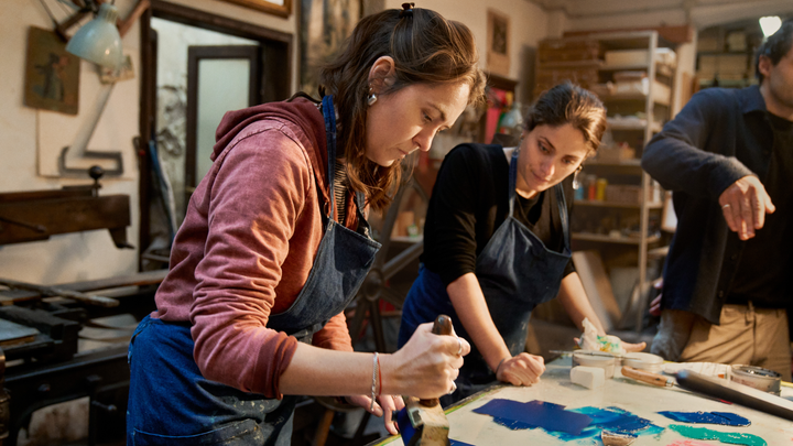 A person in a denim apron stands at a table using an ink roller to spread blue paint onto a surface. Another person watches.