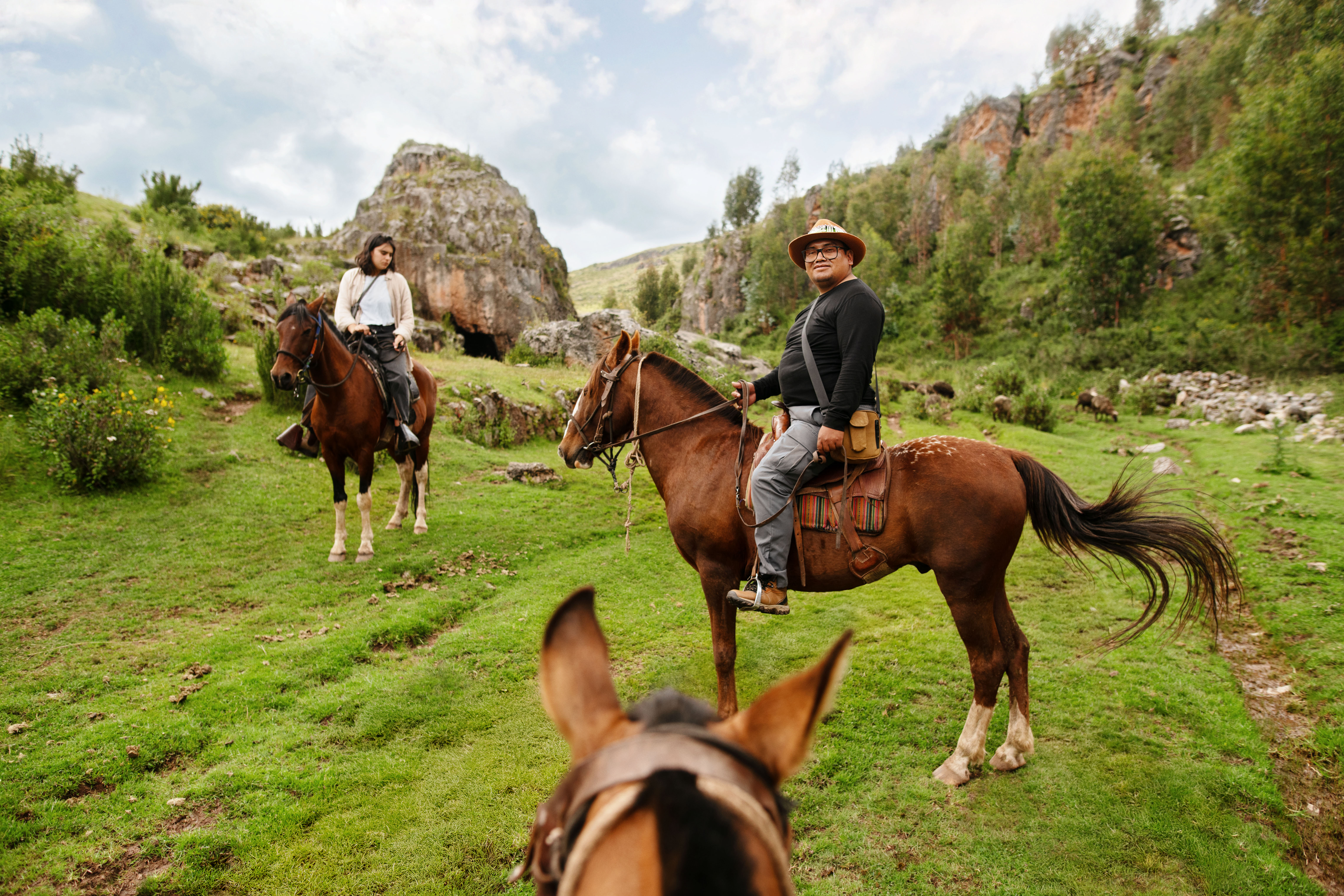 Dos personas montan a caballo en un paisaje verde y exuberante entre formaciones rocosas. La cabeza de un tercer caballo se asoma en primer plano.