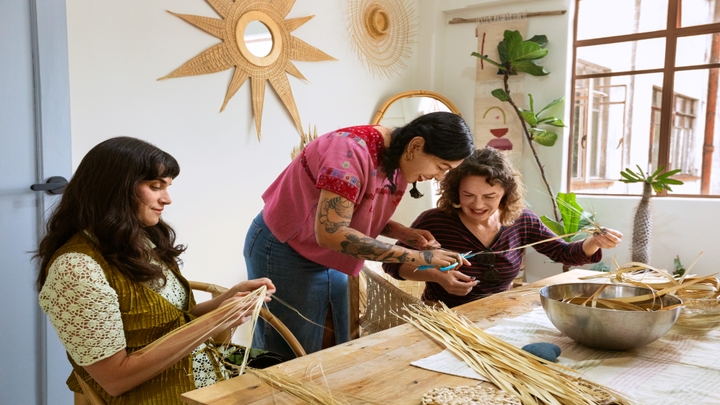 Three people gather at a wooden table and work with long, thin strips of fiber next to a window and plants.