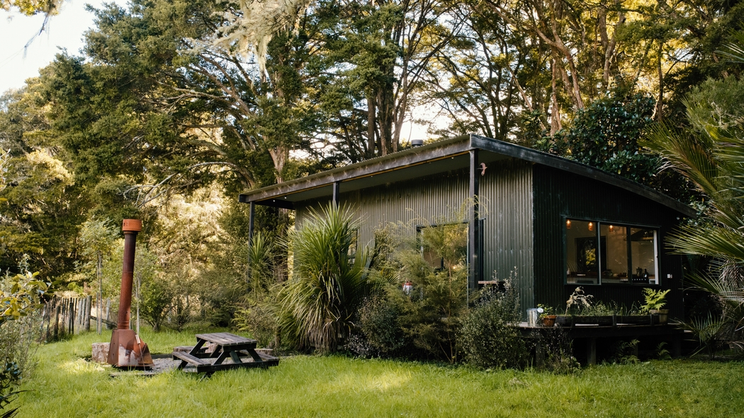 El exterior de una casa con paredes de madera oscura y grandes ventanales rodeada de árboles y plantas. Hay una mesa de pícnic al aire libre.