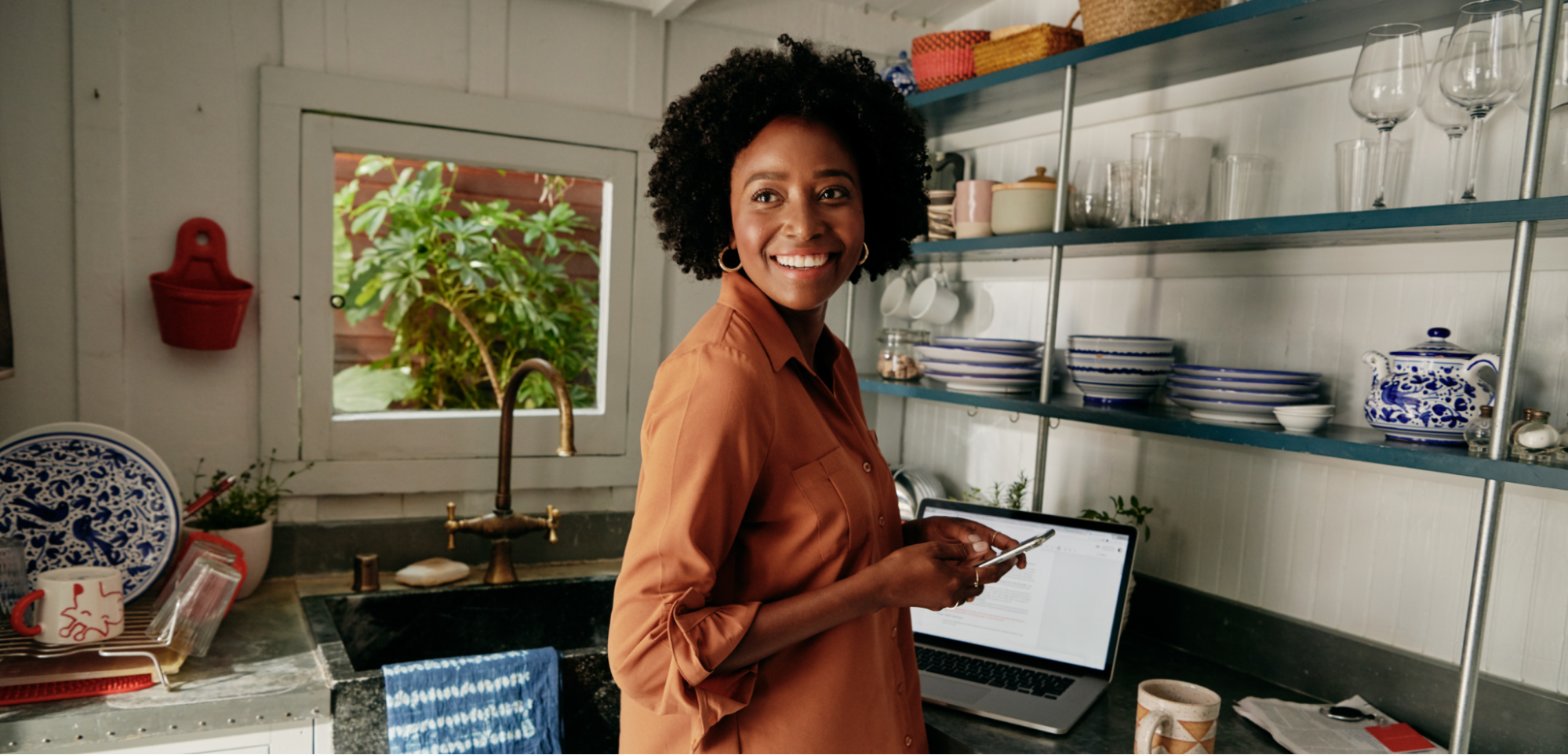 Une personne souriante, un téléphone dans la main, se tient devant un ordinateur portable ouvert sur le plan de travail d'une cuisine.