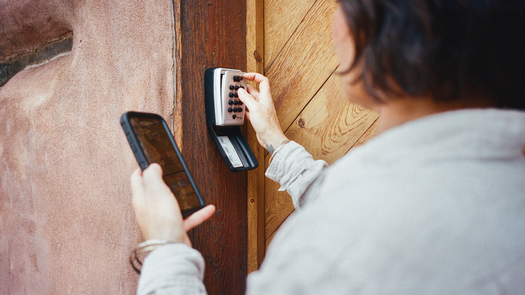 Superhost Sadie holds a mobile phone in one hand while opening a key safe that’s mounted on the wall with the other.