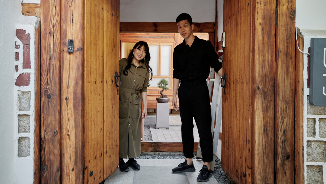 Dos anfitriones están de pie, uno a cada lado de una puerta doble de madera abierta, que deja ver un vestíbulo y un bonsái sobre un pedestal.