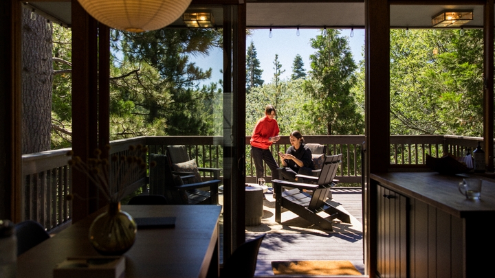 Op een terras omringd door pijnbomen zit een persoon in een Adirondack-stoel op een telefoon. Een andere persoon staat en houdt een mok vast.