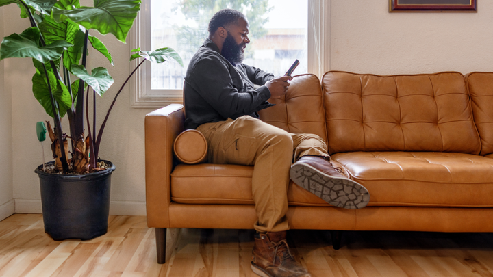 Assise sur un canapé à côté d'une fenêtre, une personne regarde son téléphone. Une grande plante Monstera se trouve à côté d'elle.