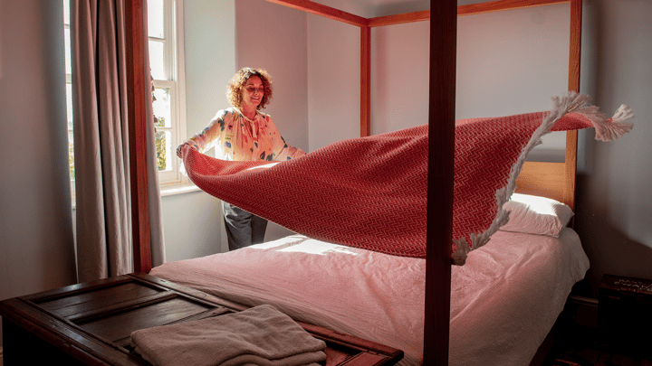 Una persona está de pie junto a una cama con dosel y tiende una colcha roja sobre sábanas blancas.