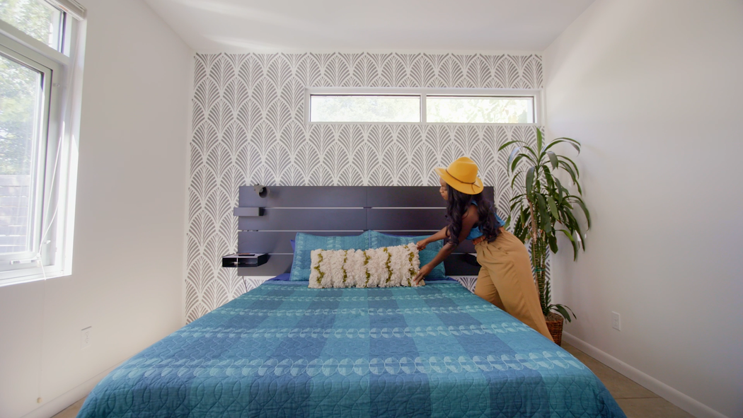 在一間明亮的房間內，一位頭戴黃色帽子的人將抱枕放在鋪著條紋與藍色花紋床單的床上。