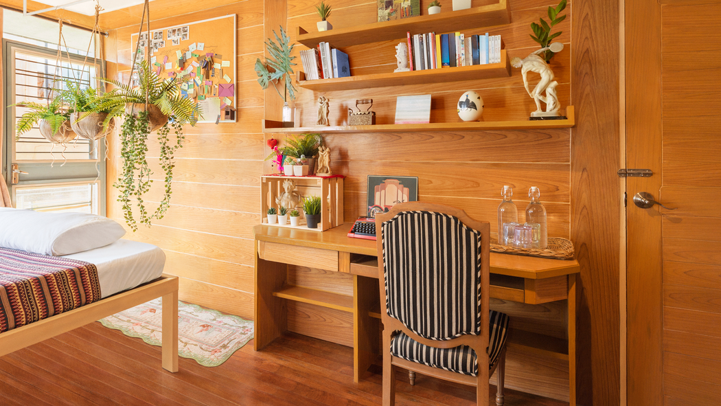 陽光明媚的臥室，窗前懸掛著藤蔓植物，木板牆壁和置物架前有一張書桌和一張椅子