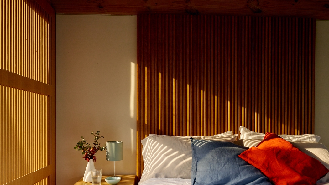 I raggi del sole illuminano una stanza vuota con cuscini di colore rosso vivo, bianco e blu disposti su un letto.
