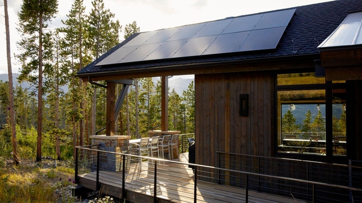 A mountain cabin surrounded by trees has picture windows, rooftop solar panels, and a wraparound deck with outdoor seating.