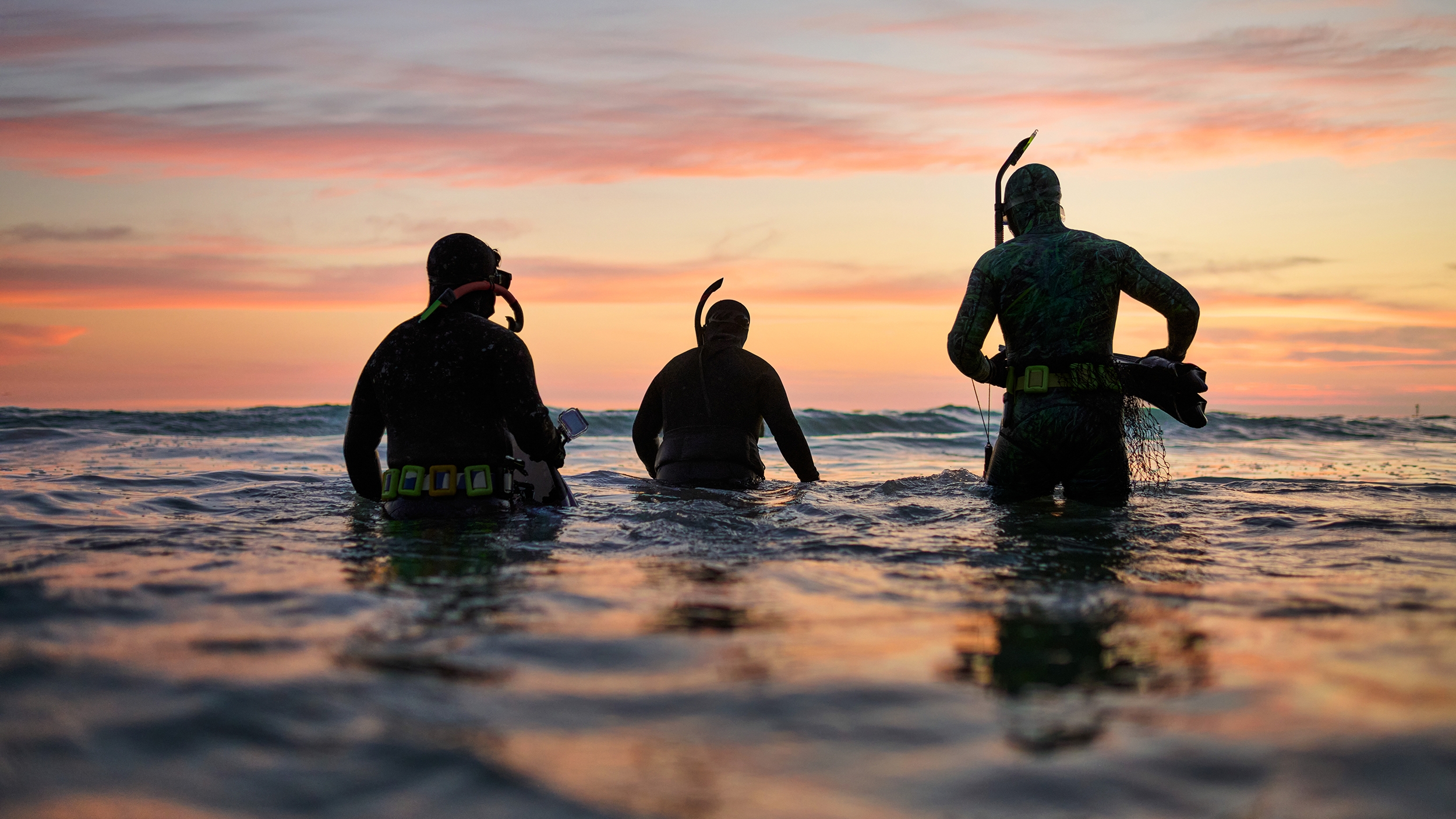 นักดำน้ำตื้น 3 คนยืนอยู่ในน้ำลึกระดับเอวช่วงพลบค่ำ มองเห็นเมฆที่เปลี่ยนเป็นสีเหลือง ชมพู และส้มหลากหลายเฉดสี