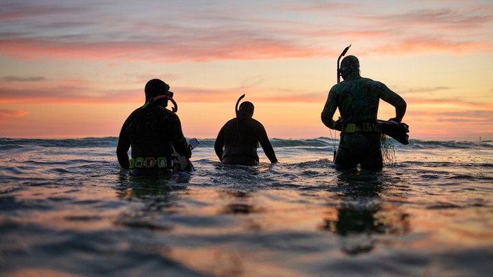 Trīs snorkelētāji stāv līdz viduklim dziļā ūdenī, krēslai izrotājot mākoņus dzeltenās, sārtās un oranžās nokrāsās.