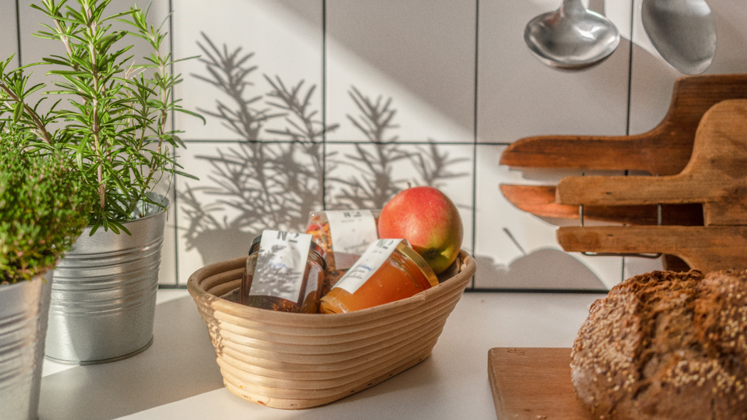 Auf einer Küchentheke befinden sich frische Kräuter, ein Korb mit Obst und Marmelade und ein Laib Brot. Dahinter hängen Servierlöffel.