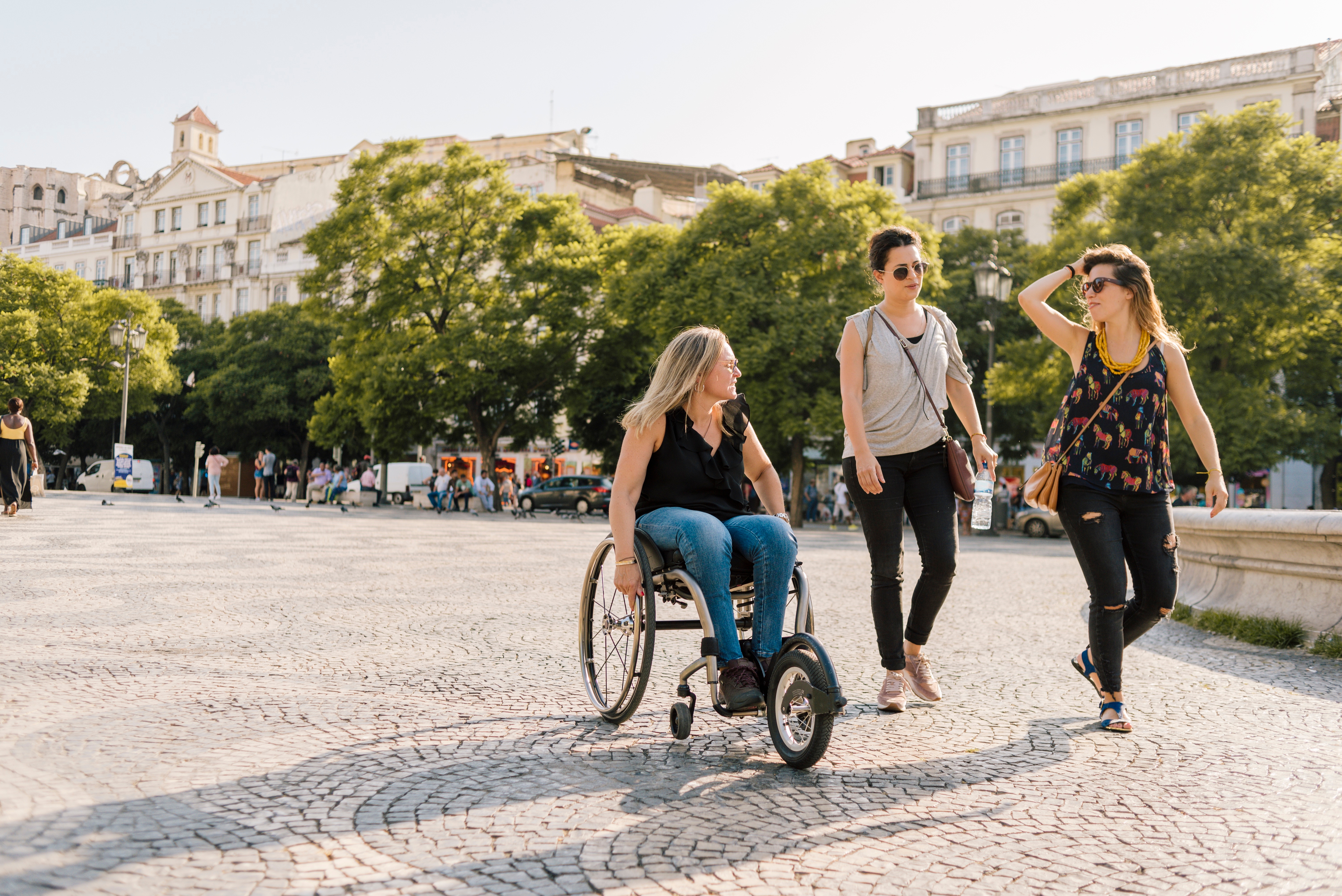 Tres personas recorren juntas una calle ancha de adoquines, bastante llana. Una de ellas va en silla de ruedas y las otras dos, caminando.