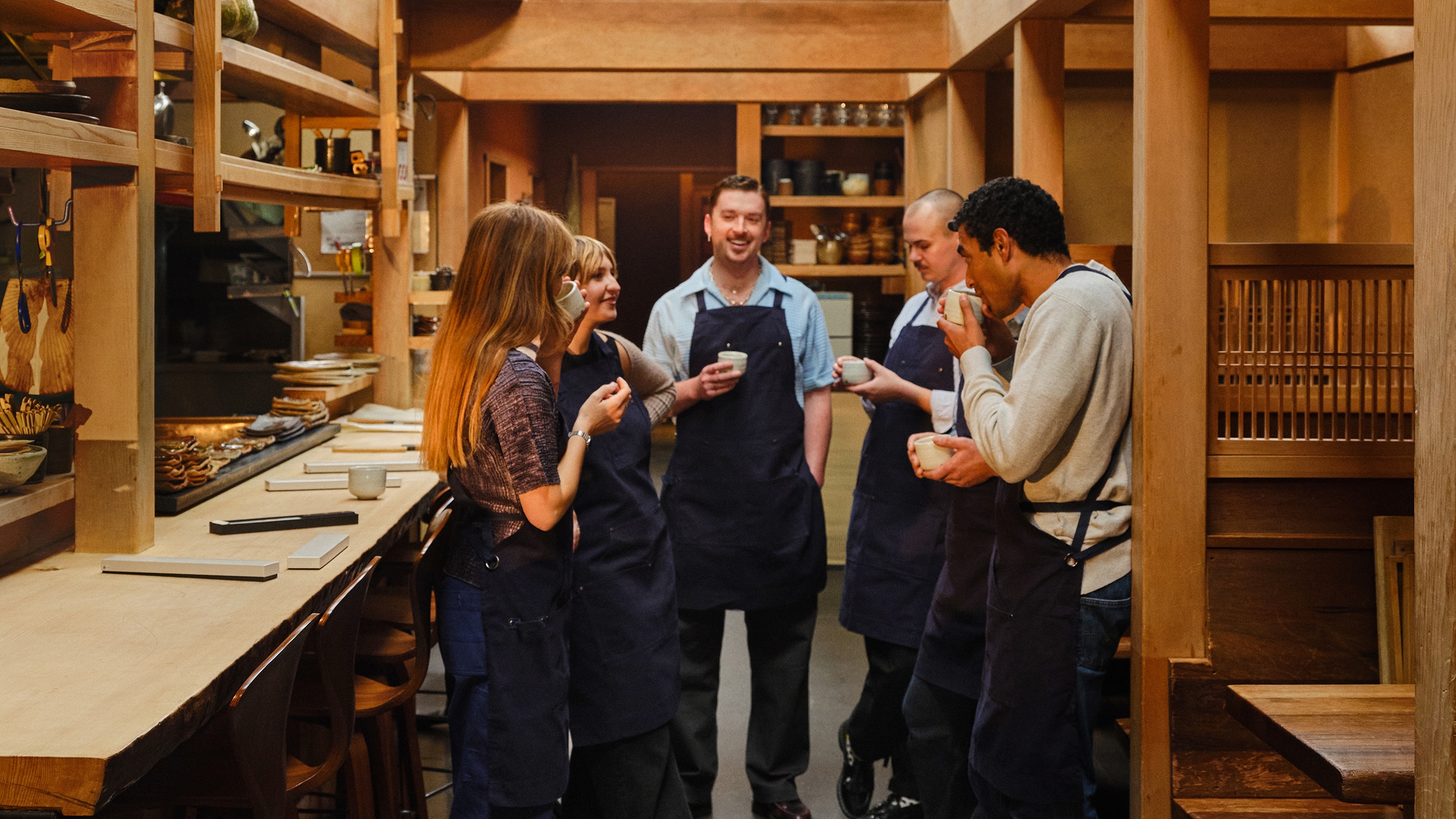 คน 6 คนสวมผ้ากันเปื้อนสีกรมท่ายืนเรียงกันเป็นครึ่งวงกลมในห้องที่กรุด้วยไม้ ทุกคนยิ้มและจิบเครื่องดื่มจากถ้วยขนาดเล็ก