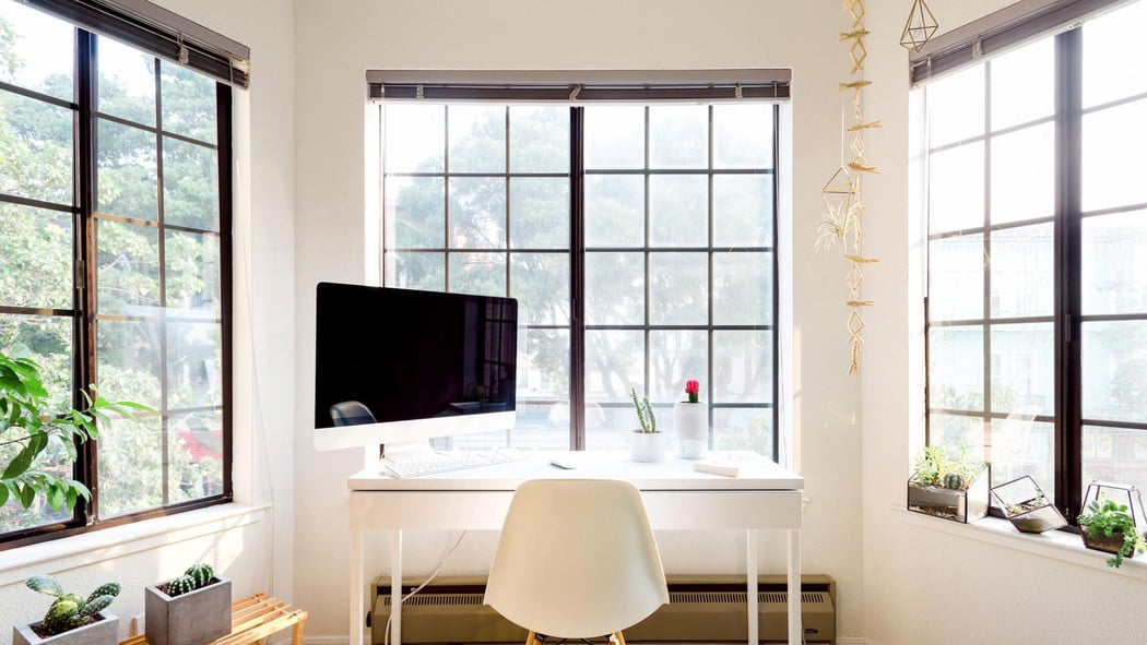 Üç büyük pencereli bir odada beyaz bir masa, sandalye ve bilgisayar monitörü bulunuyor.