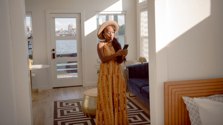 Seseorang yang mengenakan topi berwarna cokelat dan gaun panjang sedang melihat smartphone sambil berdiri di dekat sofa biru di ruang tamu yang diterangi cahaya matahari.