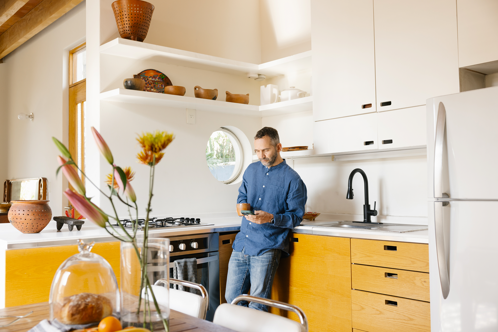 A person looks at their phone while leaning against a countertop in a kitchen with flowers on a table in the foreground.