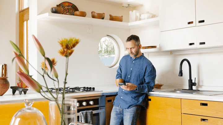A person looks at their phone while leaning against a countertop in a kitchen with flowers on a table in the foreground.
