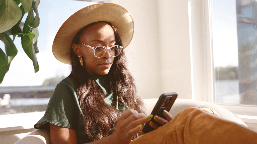 A person wearing glasses and a wide-brimmed hat sits in a beige chair in the sunlight while looking at a smartphone.