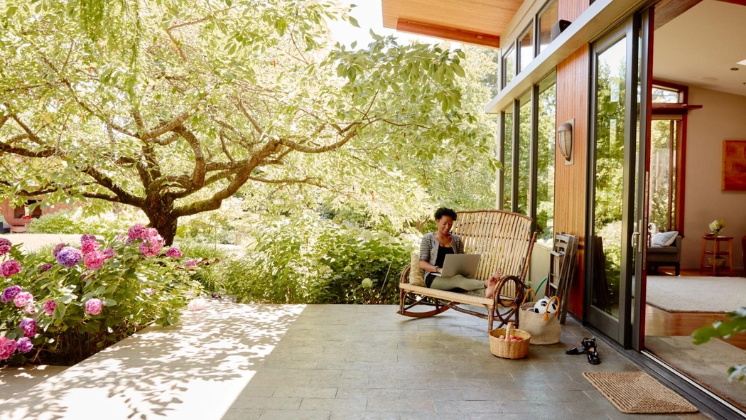 一位身穿緊身褲和開襟毛衣的人坐在室外露台上用筆電打字，四周圍繞著樹木和花朵。