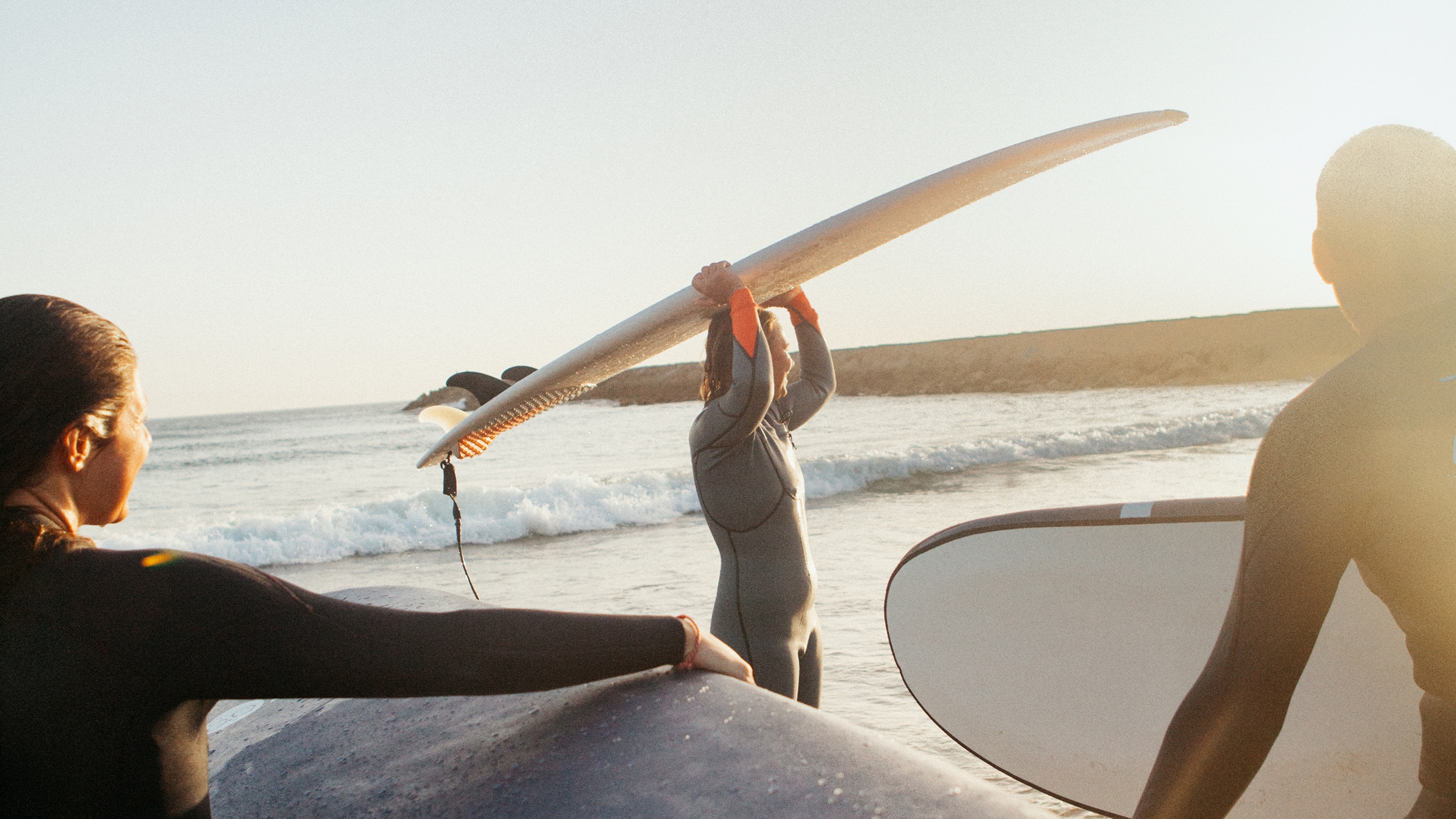 นักโต้คลื่นสวมเว็ทสูทยืนในน้ำตื้นแบกกระดานโต้คลื่นบนหัว อีกสองคนหนีบกระดานโต้คลื่นใต้แขน