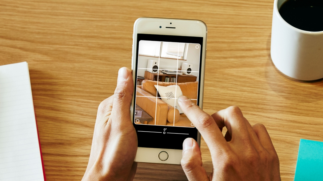 Duas mãos seguram um telefone e editam uma imagem de uma sala de estar, que tem um sofá de couro e uma mesa de apoio de madeira abaixo de uma janela.