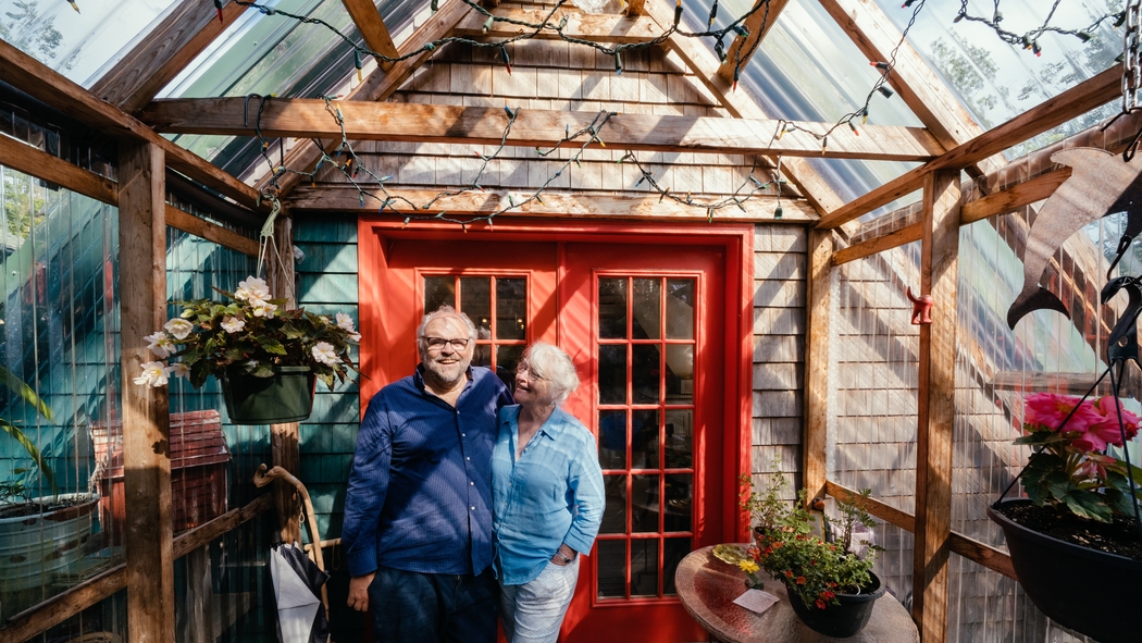 Deux hôtes sourient et se tiennent bras dessus bras dessous devant un chalet en A avec des bardeaux de bois naturel et une porte à deux battants rouge.