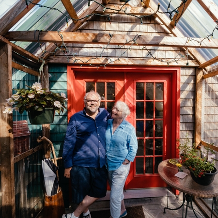 Dva hostitelé se usmívají a stojí ruku v ruce před domovem ve tvaru A s šindelemi z přírodního dřeva a červenými francouzskými dveřmi.