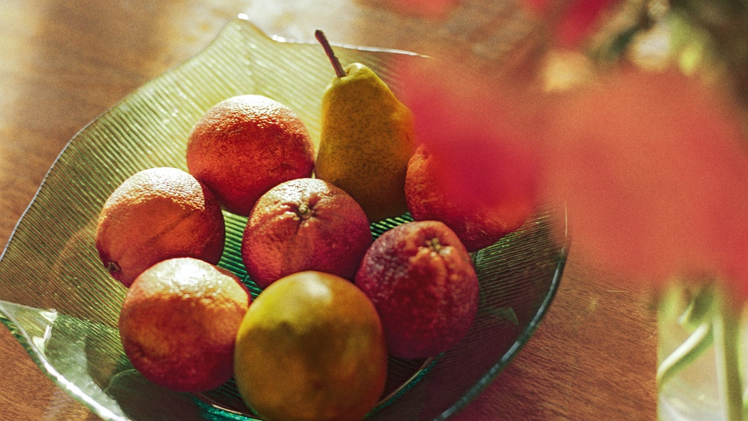 Vers fruit, waaronder citrusvruchten en peren, in een groene kom op een houten tafel naast een vaas met bloemen.