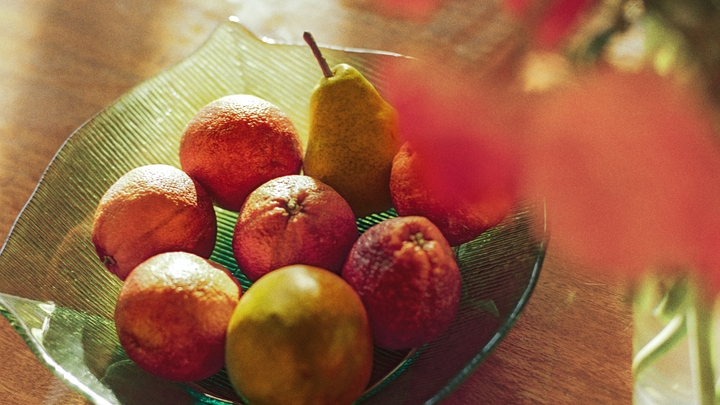 Vers fruit, waaronder citrusvruchten en peren, in een groene kom op een houten tafel naast een vaas met bloemen.