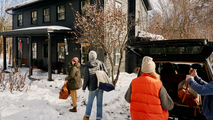 Four adults in hats and coats unload bags out of a car boot and carry them toward a snow-covered house.