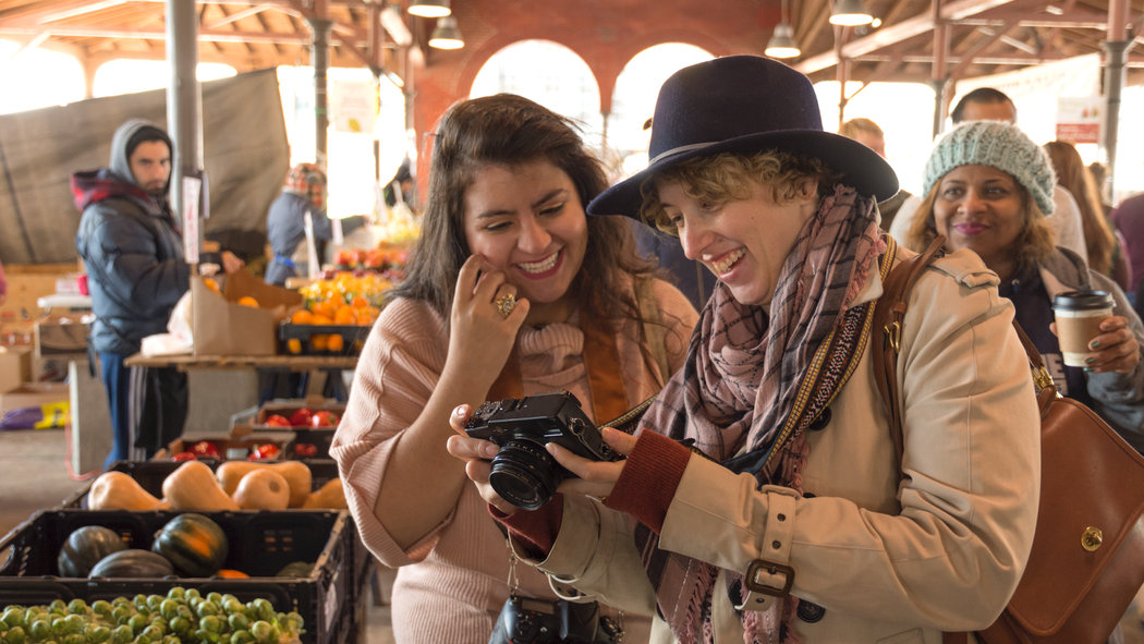 Anfitrião e hóspede em um mercado de agricultores, sorrindo e olhando para uma imagem na tela de uma câmera digital